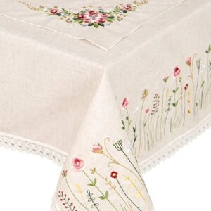 May include: Cream-colored linen tablecloth with embroidered floral details. Features a corner design with pink, red, and yellow flowers, and a border of delicate white lace. The tablecloth is draped over a wooden table.