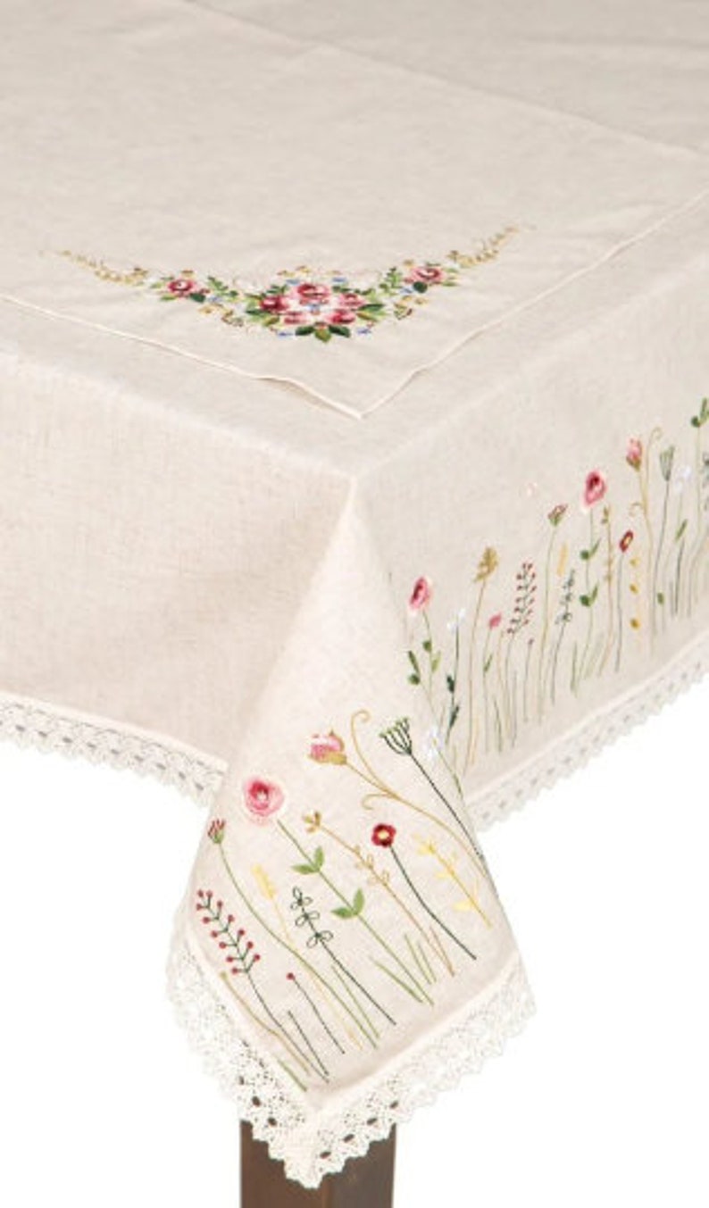May include: Cream-colored linen tablecloth with embroidered floral details. Features a corner design with pink, red, and yellow flowers, and a border of delicate white lace. The tablecloth is draped over a wooden table.