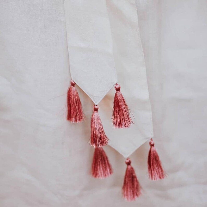 May include: Close-up of white fabric with a decorative design. The design features a series of pink tassels hanging from a white, triangular-shaped fabric. The tassels are a deep pink colour and add a touch of elegance to the design.