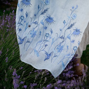 Puede incluir: Tela de lino azul claro con dise&ntilde;o floral bordado en azul. El bordado presenta flores y tallos detallados en diferentes tonos de azul. La tela est&aacute; drapeada sobre un fondo de plantas de lavanda.