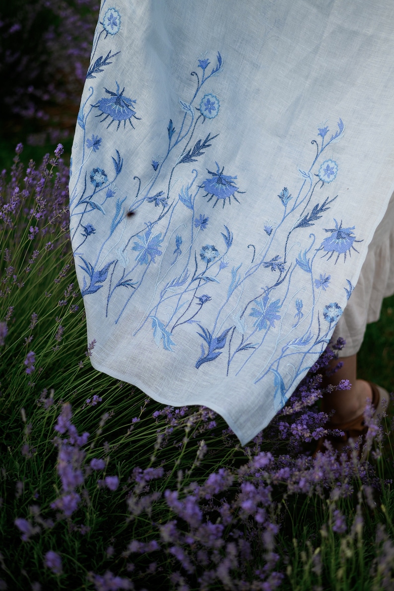 Puede incluir: Tela de lino azul claro con dise&ntilde;o floral bordado en azul. El bordado presenta flores y tallos detallados en diferentes tonos de azul. La tela est&aacute; drapeada sobre un fondo de plantas de lavanda.