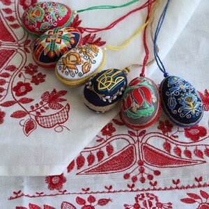 K&ouml;nnte beinhalten: F&uuml;nf bestickte Ostereier mit bunten Blumenmustern und geometrischen Designs. Die Eier h&auml;ngen an Schn&uuml;ren und werden auf einer wei&szlig;en Leinen-Tischdecke mit roten Stickereien pr&auml;sentiert.