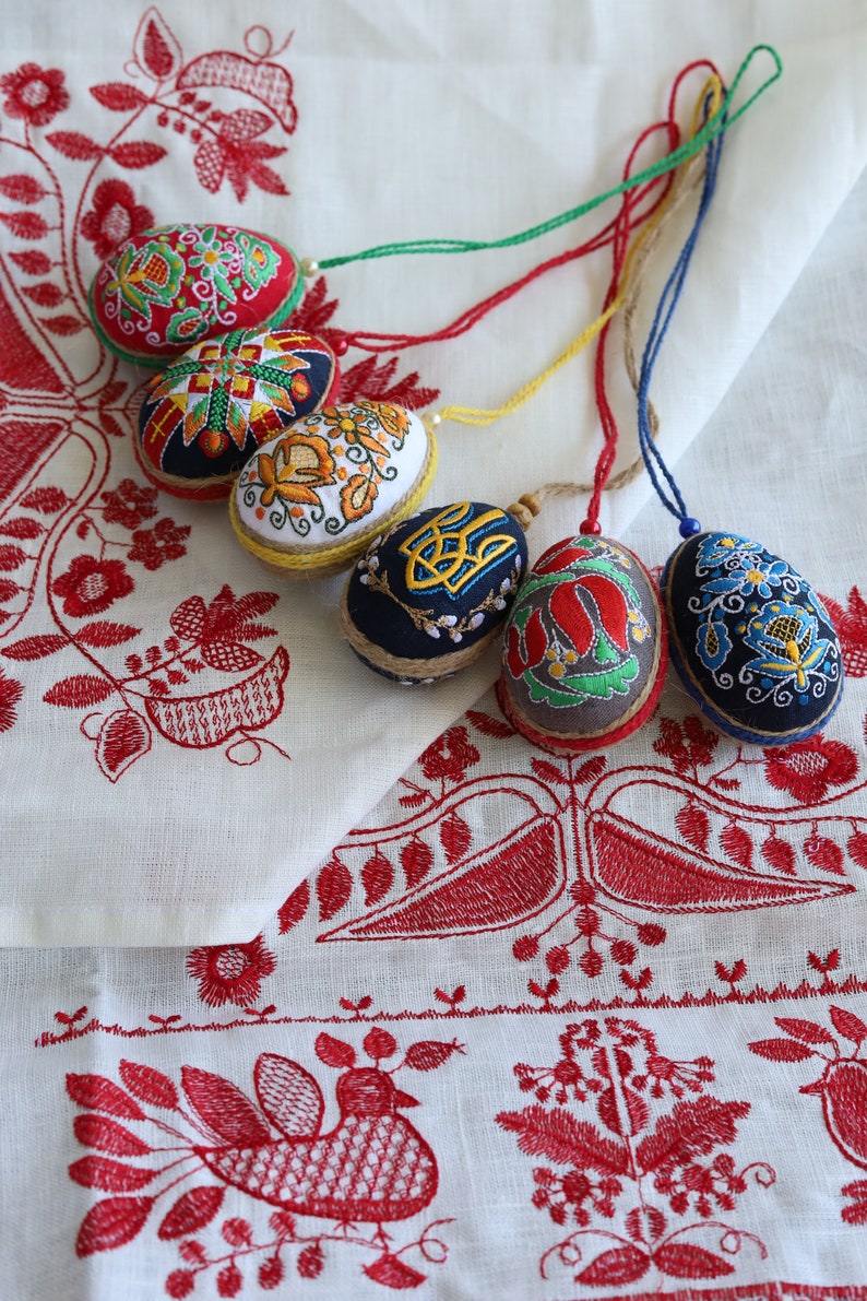 K&ouml;nnte beinhalten: F&uuml;nf bestickte Ostereier mit bunten Blumenmustern und geometrischen Designs. Die Eier h&auml;ngen an Schn&uuml;ren und werden auf einer wei&szlig;en Leinen-Tischdecke mit roten Stickereien pr&auml;sentiert.