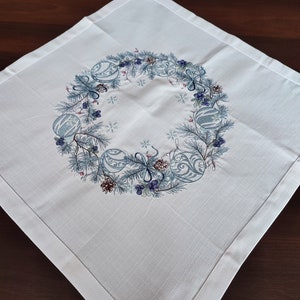 K&ouml;nnte beinhalten: Quadratische wei&szlig;e Tischdecke mit einem bestickten Kranzdesign. Der Kranz zeigt blaue Ornamente, Tannennadeln, Beeren und Tannenzapfen. Die Tischdecke hat ein subtiles strukturiertes Muster und einen fertigen Rand, f&uuml;r Weihnachten.