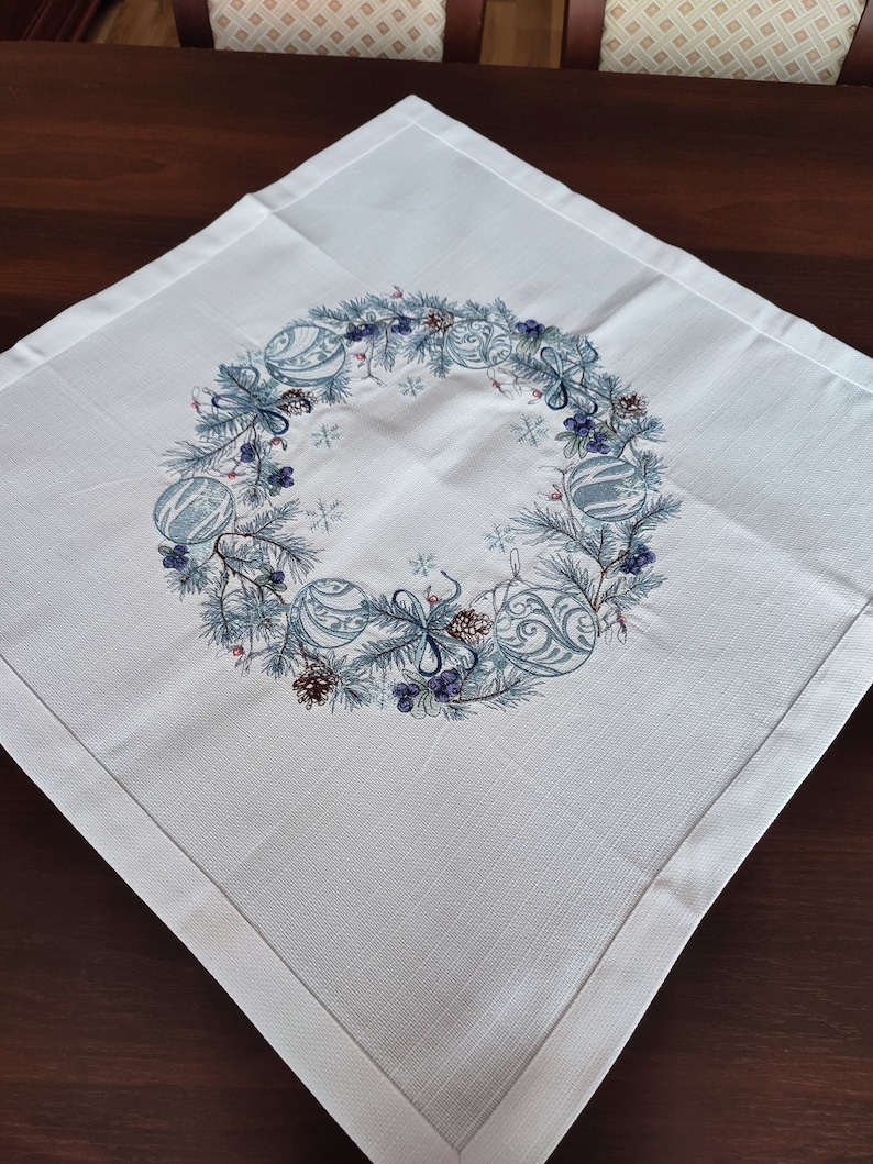 K&ouml;nnte beinhalten: Quadratische wei&szlig;e Tischdecke mit einem bestickten Kranzdesign. Der Kranz zeigt blaue Ornamente, Tannennadeln, Beeren und Tannenzapfen. Die Tischdecke hat ein subtiles strukturiertes Muster und einen fertigen Rand, f&uuml;r Weihnachten.