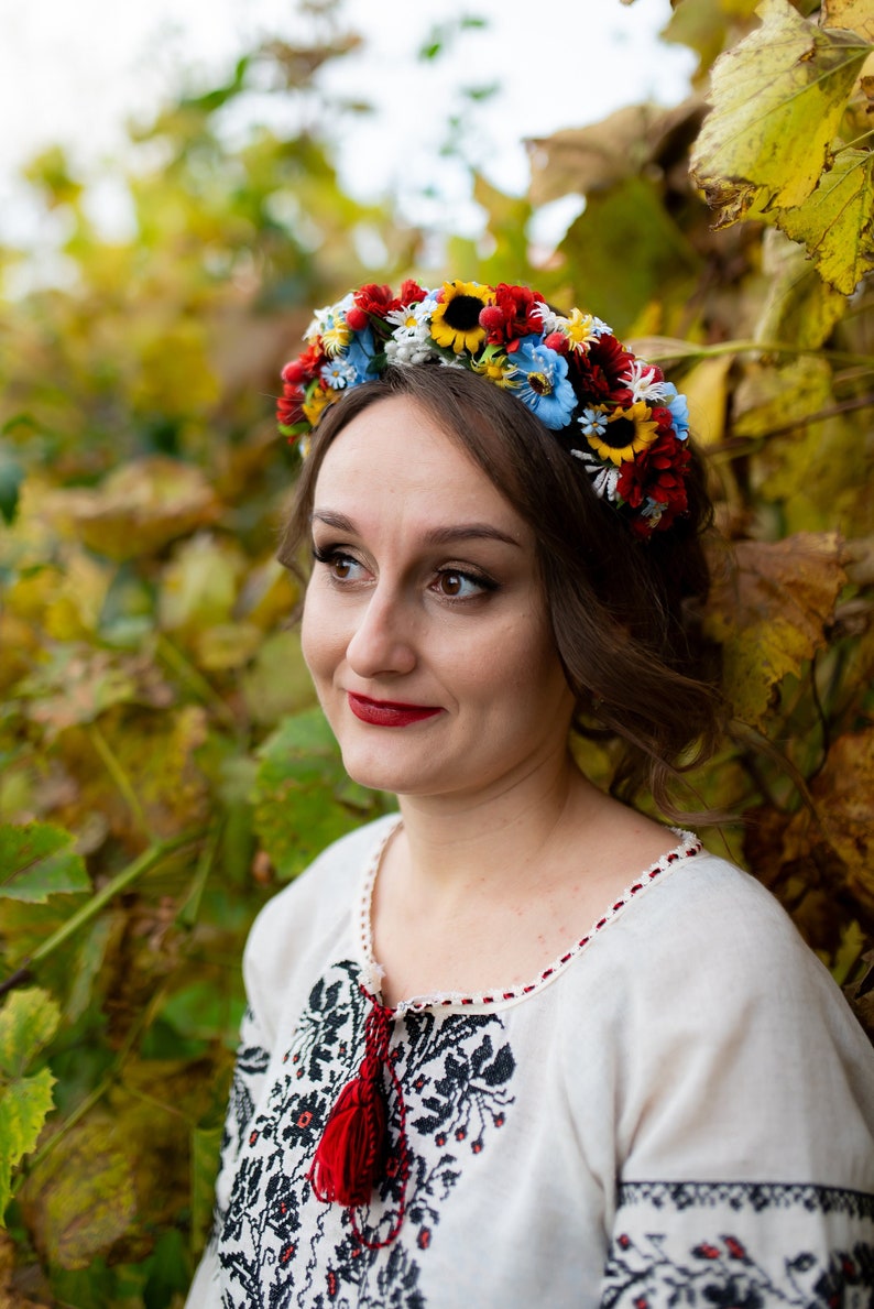 Pode incluir: Uma mulher vestindo uma blusa branca bordada com uma borla vermelha e uma coroa de flores na cabe&ccedil;a. A coroa &eacute; feita de flores vermelhas, amarelas, azuis e brancas.