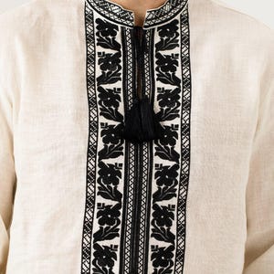 May include: Cream-colored linen shirt with intricate black floral embroidery around the collar and down the front. The shirt features a mandarin collar and a black tassel detail at the neckline. The embroidery is detailed and creates a striking contrast against the light fabric.