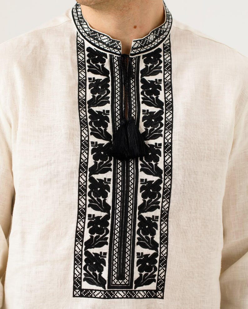 May include: Cream-colored linen shirt with intricate black floral embroidery around the collar and down the front. The shirt features a mandarin collar and a black tassel detail at the neckline. The embroidery is detailed and creates a striking contrast against the light fabric.