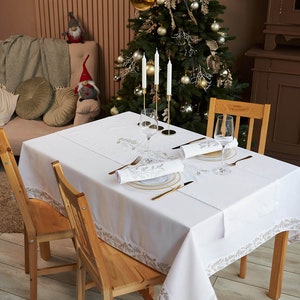 Puede incluir: Un mantel blanco con un borde de hojas doradas est&aacute; puesto para una comida festiva. La mesa est&aacute; puesta con platos, servilletas y cubiertos con borde dorado. Tres velas blancas est&aacute;n en un candelabro dorado. Dos sillas de madera est&aacute;n en la mesa.