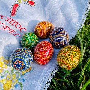 K&ouml;nnte beinhalten: Eine Sammlung von kunstvoll dekorierten Ostereiern in Rot, Blau, Gelb, Gr&uuml;n und Braun. Die Eier sind auf einem wei&szlig;en Tuch mit Stickereien platziert und von gr&uuml;nem Gras umgeben.