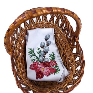 May include: A white fabric with red and green embroidered flowers and a sprig of willow branches is inside a brown wicker basket.