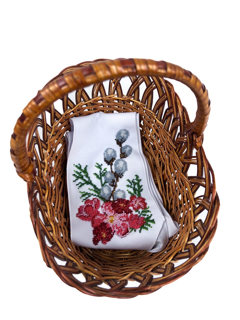 May include: A white fabric with red and green embroidered flowers and a sprig of willow branches is inside a brown wicker basket.