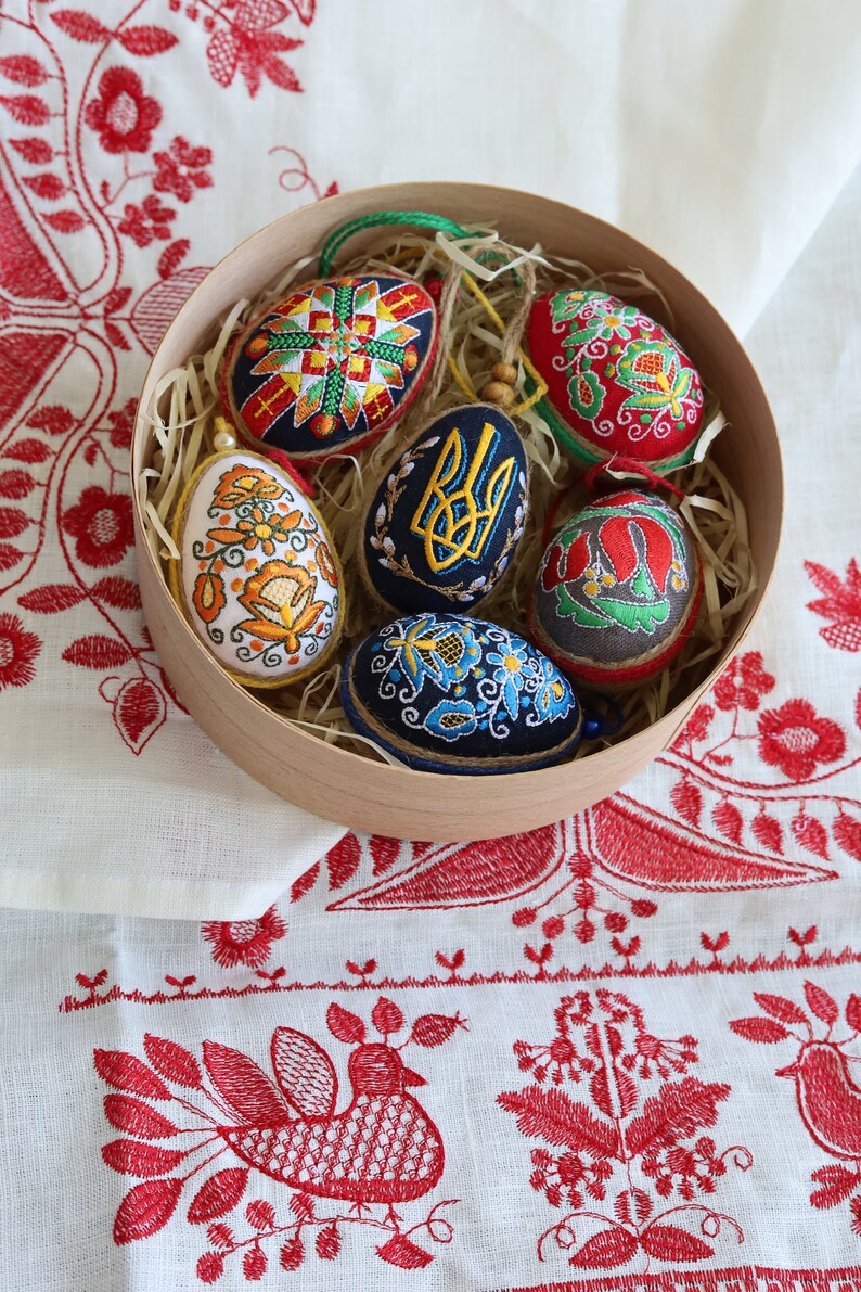 K&ouml;nnte beinhalten: Sechs bestickte Ostereier in einer Holzkiste mit Stroh. Die Eier sind mit traditionellen ukrainischen Volksmotiven verziert, darunter Blumen, geometrische Muster und der ukrainische Dreizack.