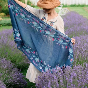 May include: A woman wearing a straw hat and a white dress with embroidery stands in a field of lavender. She is holding a blue scarf with floral embroidery.