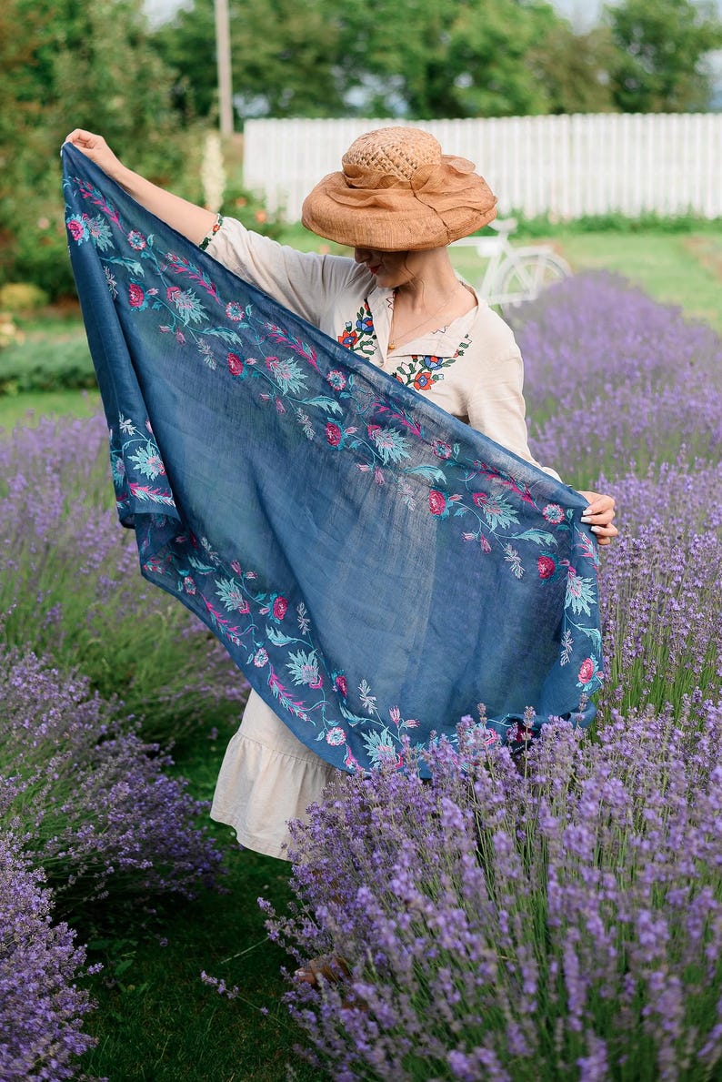 May include: A woman wearing a straw hat and a white dress with embroidery stands in a field of lavender. She is holding a blue scarf with floral embroidery.