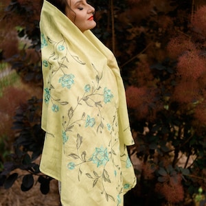 May include: A woman wearing a yellow linen scarf with a floral embroidery design. The scarf has a light blue floral pattern on a yellow background.