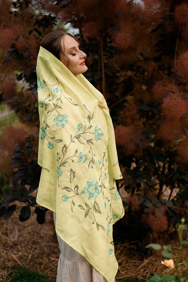 May include: A woman wearing a yellow linen scarf with a floral embroidery design. The scarf has a light blue floral pattern on a yellow background.