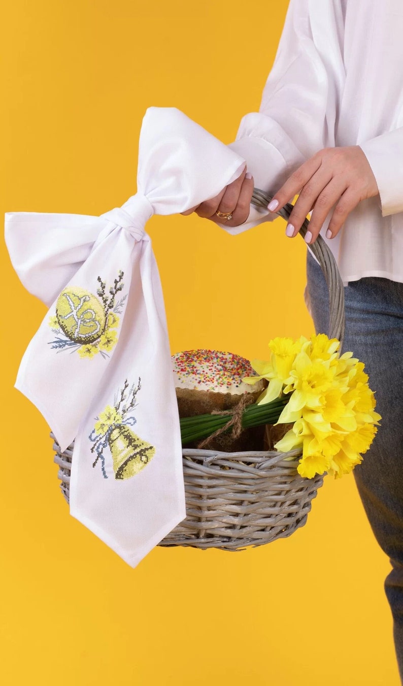 K&ouml;nnte beinhalten: Ein Weidenkorb gef&uuml;llt mit einem gelben Kuchen, Narzissen und gr&uuml;nen Zweigen, mit einer wei&szlig;en bestickten Stoffserviette mit einem Muster aus gelbem Osterei und Glocke.