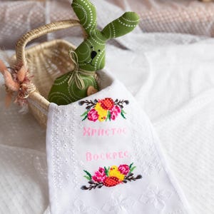 May include: A white embroidered hand towel with a pink embroidered message that says "ХРИСТОС ВОСКРЕС" and a floral design with yellow chicks. A green bunny is in a wicker basket in the background.