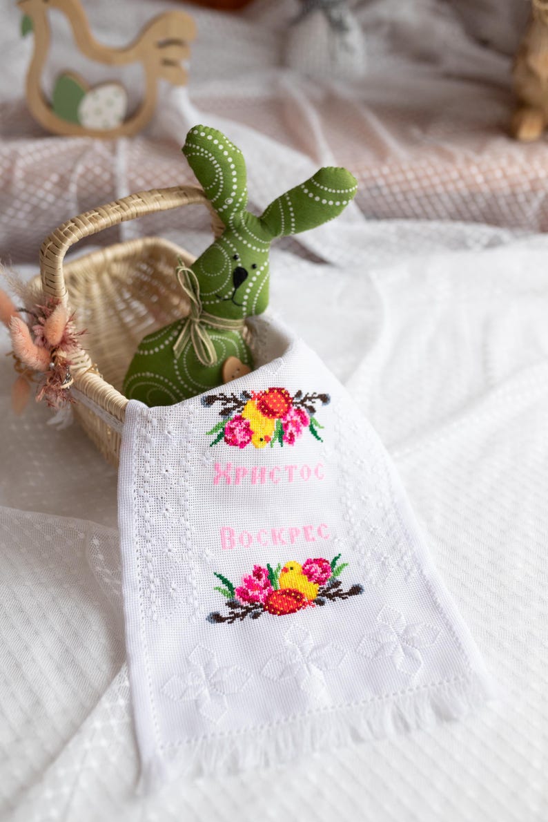 May include: A white embroidered hand towel with a pink embroidered message that says "ХРИСТОС ВОСКРЕС" and a floral design with yellow chicks. A green bunny is in a wicker basket in the background.