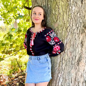 Puede incluir: Una mujer lleva una blusa negra bordada con dise&ntilde;os florales rojos y blancos, combinada con una falda vaquera azul claro. Est&aacute; de pie junto a un tronco de &aacute;rbol grande.