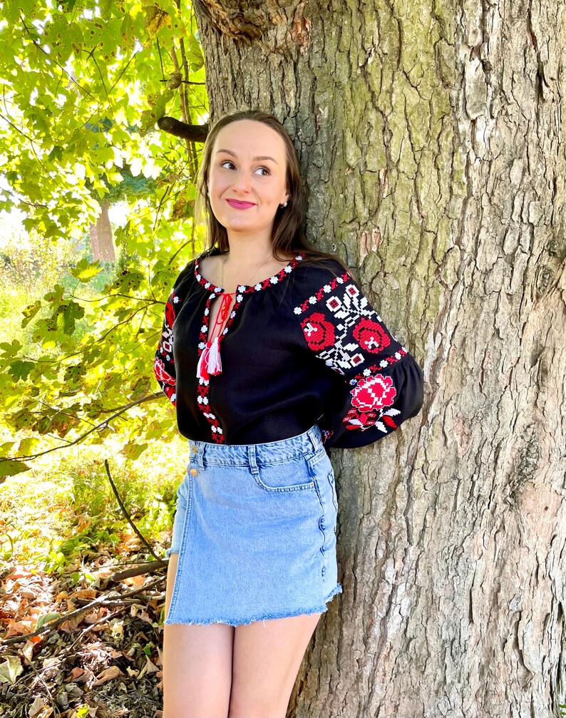 Puede incluir: Una mujer lleva una blusa negra bordada con dise&ntilde;os florales rojos y blancos, combinada con una falda vaquera azul claro. Est&aacute; de pie junto a un tronco de &aacute;rbol grande.