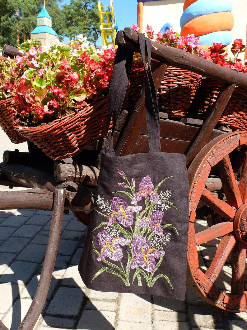 May include: A black tote bag with embroidered purple iris flowers on a dark background. The bag is hanging on a wooden cart with a red wheel.