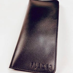 May include: A rectangular black wallet with rounded corners and visible stitching along the edges. The word FLAXSI is embossed in capital letters on the bottom right corner of the front surface. The wallet has a smooth, matte finish throughout.