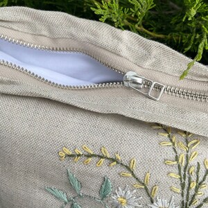 May include: A beige linen pillow with a zipper closure and embroidered floral design featuring white daisies and yellow flowers.