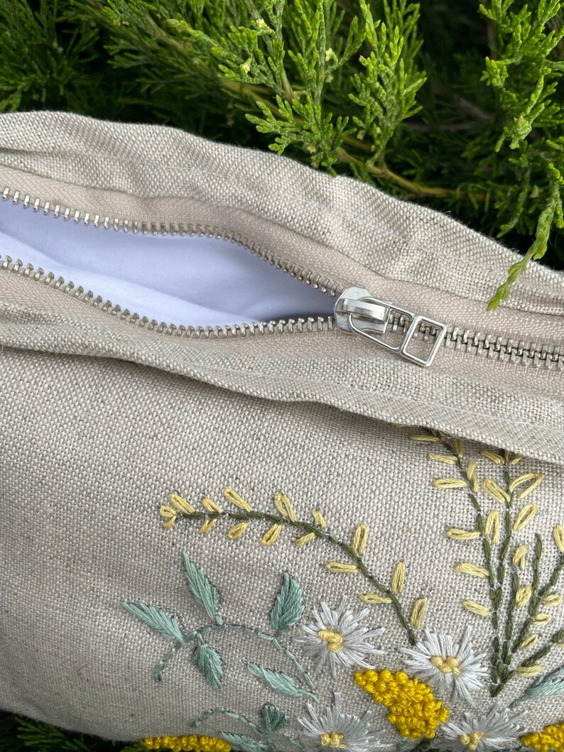 May include: A beige linen pillow with a zipper closure and embroidered floral design featuring white daisies and yellow flowers.