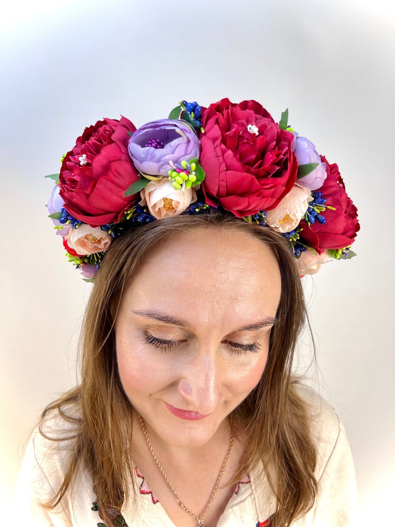 May include: A red, pink, and purple flower crown with green leaves and small blue berries. The crown is worn by a person with brown hair.