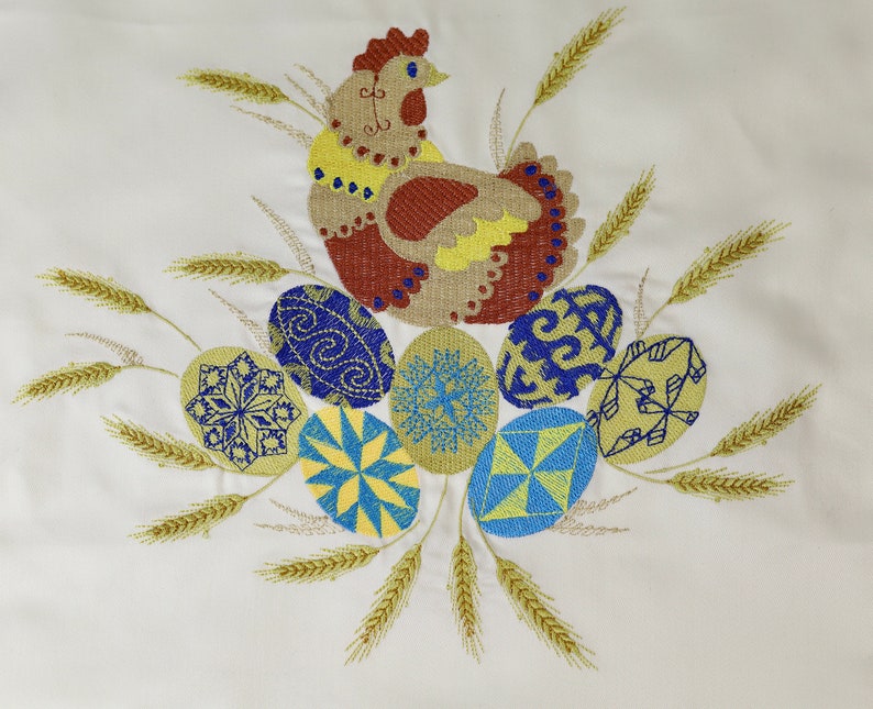 Pu&ograve; includere: Disegno ricamato di una gallina marrone e gialla circondata da spighe di grano e uova colorate con motivi blu, gialli e bianchi.