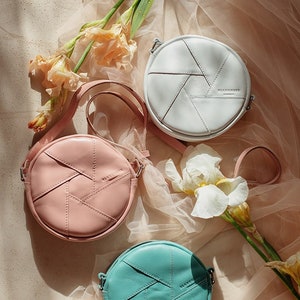 May include: Three round leather crossbody bags in white, pink, and teal. Each bag has a geometric design and a small logo on the front.