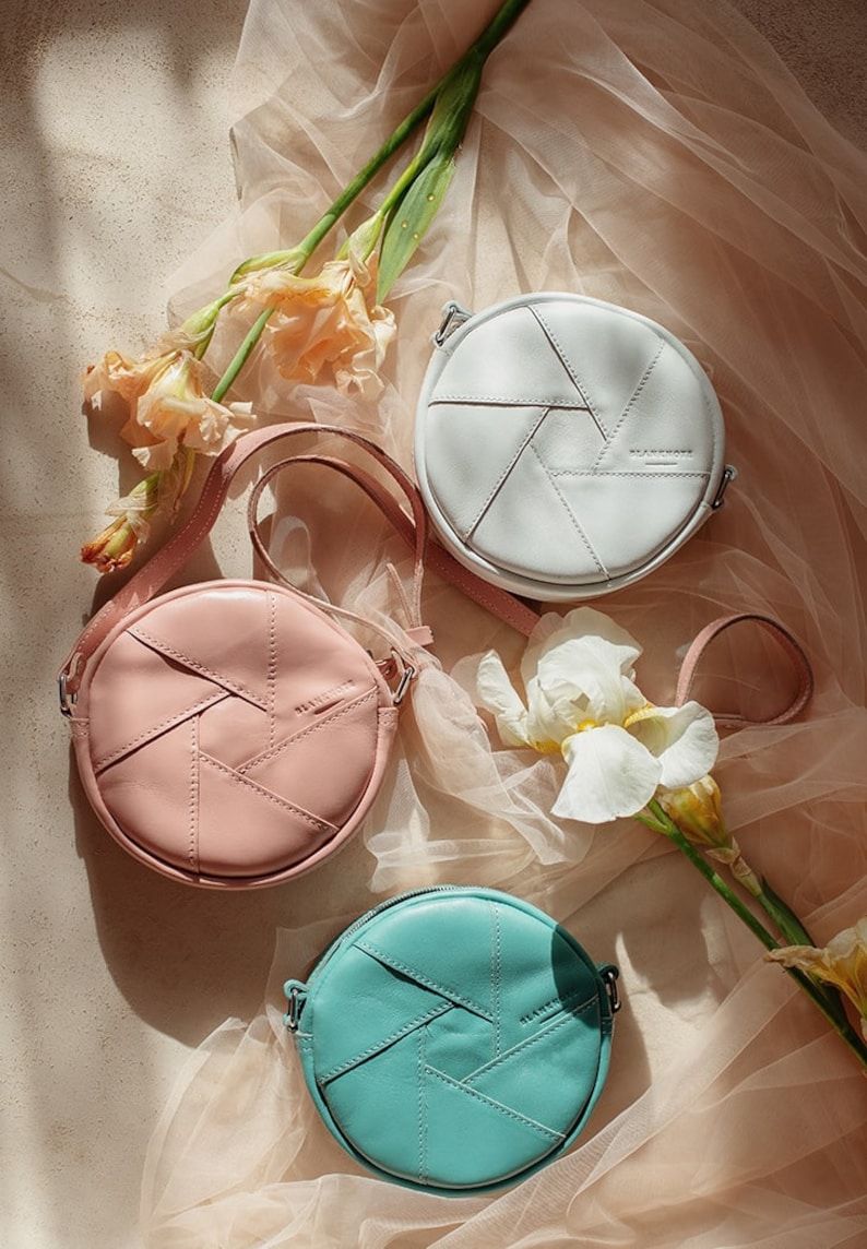 May include: Three round leather crossbody bags in white, pink, and teal. Each bag has a geometric design and a small logo on the front.