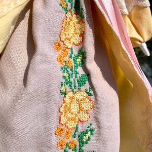 Puede incluir: Una mu&ntilde;eca vintage con un vestido rosa claro y amarillo. El vestido tiene un dise&ntilde;o floral bordado en la falda con hojas verdes y flores amarillas.