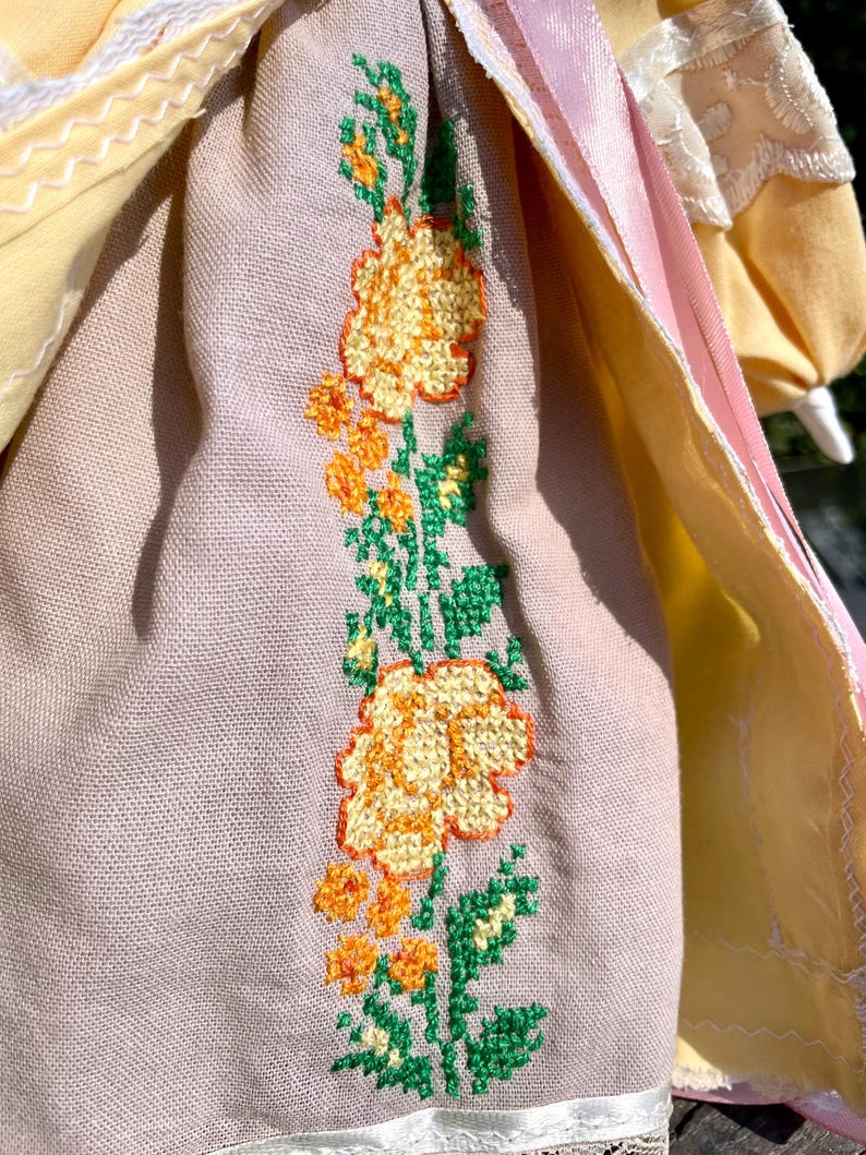Puede incluir: Una mu&ntilde;eca vintage con un vestido rosa claro y amarillo. El vestido tiene un dise&ntilde;o floral bordado en la falda con hojas verdes y flores amarillas.