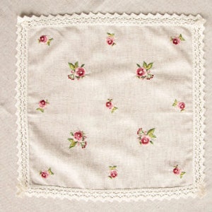 May include: A square, beige linen handkerchief with embroidered pink roses and green leaves. The handkerchief is trimmed with a white lace border. The background is a beige linen surface.