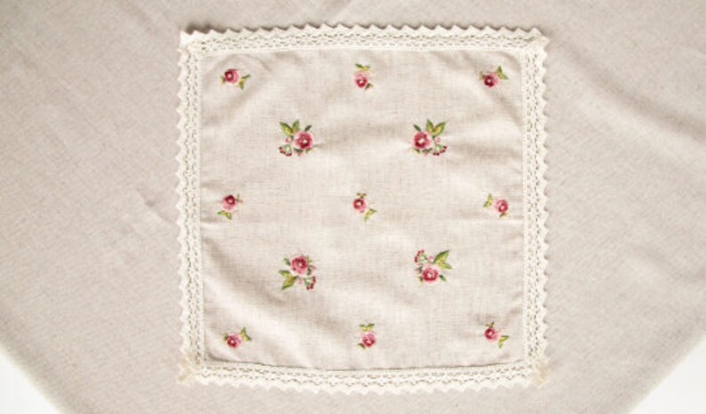 May include: A square, beige linen handkerchief with embroidered pink roses and green leaves. The handkerchief is trimmed with a white lace border. The background is a beige linen surface.