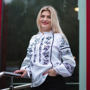 Puede incluir: Una blusa blanca con mangas largas y un intrincado bordado floral azul oscuro. La blusa presenta un cuello con volantes y se combina con pantalones negros. Las mangas tienen un patr&oacute;n bordado detallado cerca de los pu&ntilde;os.