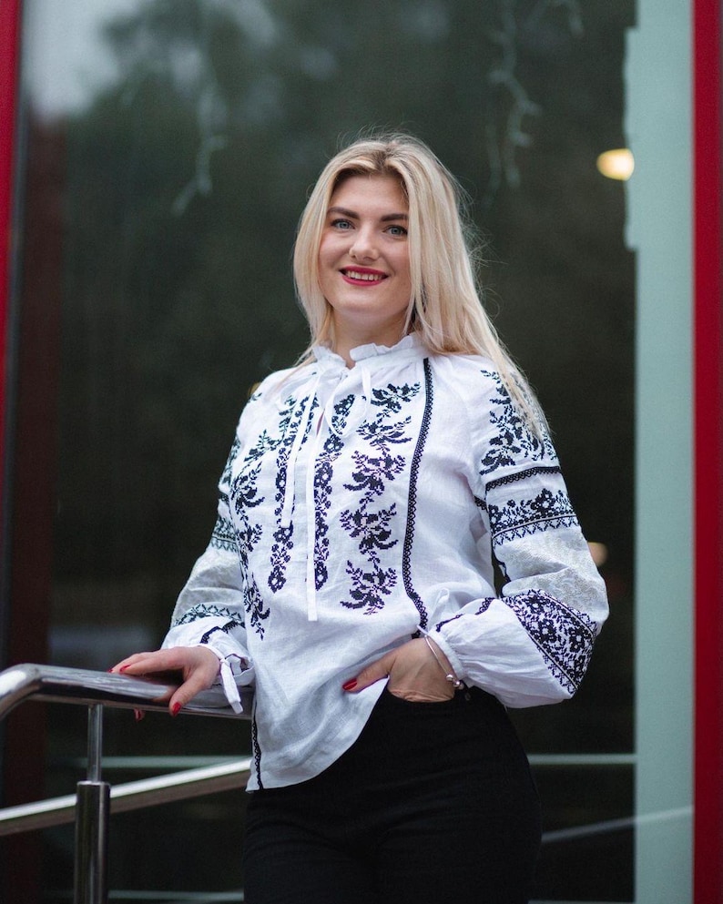 Puede incluir: Una blusa blanca con mangas largas y un intrincado bordado floral azul oscuro. La blusa presenta un cuello con volantes y se combina con pantalones negros. Las mangas tienen un patr&oacute;n bordado detallado cerca de los pu&ntilde;os.
