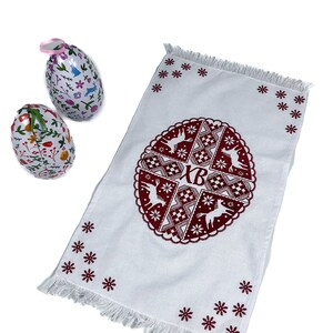 May include: White Easter towel with a burgundy circular design featuring deer, geometric patterns, and the letters "XB." Two decorative Easter eggs with floral and bunny designs are also present. The towel has a fringed edge and star-shaped embroidery.