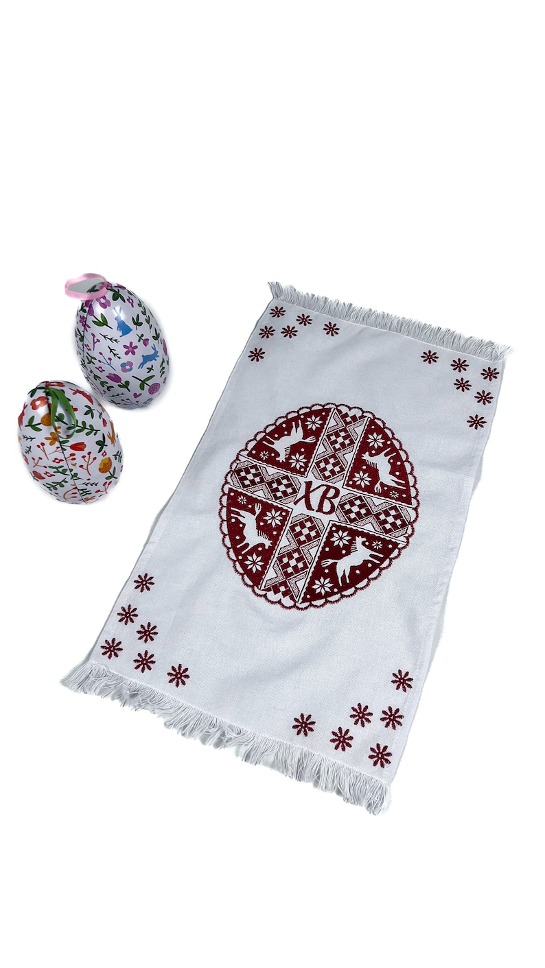 May include: White Easter towel with a burgundy circular design featuring deer, geometric patterns, and the letters "XB." Two decorative Easter eggs with floral and bunny designs are also present. The towel has a fringed edge and star-shaped embroidery.