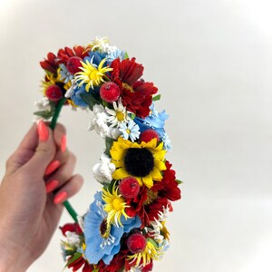 Pode incluir: Uma faixa de cabe&ccedil;a floral com flores vermelhas, amarelas, azuis e brancas. A faixa de cabe&ccedil;a &eacute; feita de arame verde e tem um girassol no centro.