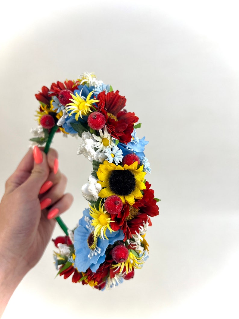 Pode incluir: Uma faixa de cabe&ccedil;a floral com flores vermelhas, amarelas, azuis e brancas. A faixa de cabe&ccedil;a &eacute; feita de arame verde e tem um girassol no centro.