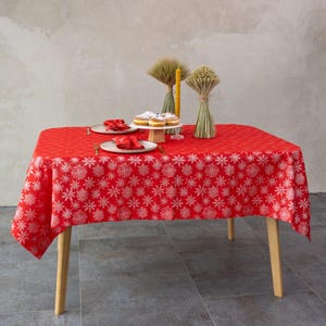 May include: A red rectangular tablecloth with a white snowflake pattern covers a wooden table. Two plates with red napkins and gold forks are set. A cake stand holds donuts. Decorative wheat bundles and a candle complete the festive scene.