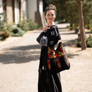 May include: Black dress with white embroidered details and a matching black handbag. The handbag features colorful floral embroidery in red, yellow, and orange. The dress has long sleeves with white geometric patterns.