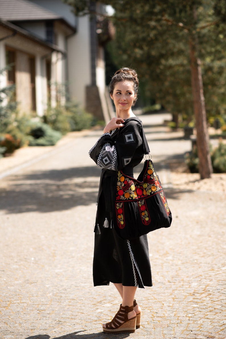 May include: Black dress with white embroidered details and a matching black handbag. The handbag features colorful floral embroidery in red, yellow, and orange. The dress has long sleeves with white geometric patterns.