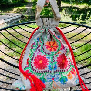 May include: A small, gray linen drawstring bag with red tassels. The bag is embroidered with colorful flowers and leaves.