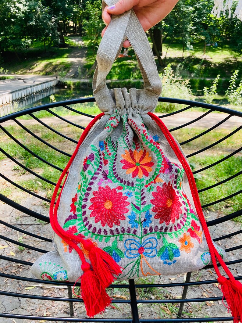 May include: A small, gray linen drawstring bag with red tassels. The bag is embroidered with colorful flowers and leaves.