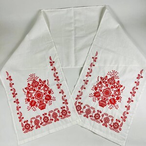 May include: White table runner with red embroidered floral designs. The design features two bouquets of flowers in a vase with leaves and stems.